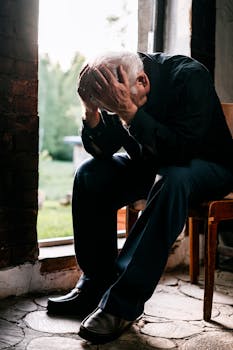 Senior man in black, grieving alone, sitting on a wooden chair by a doorway in a pensive pose.